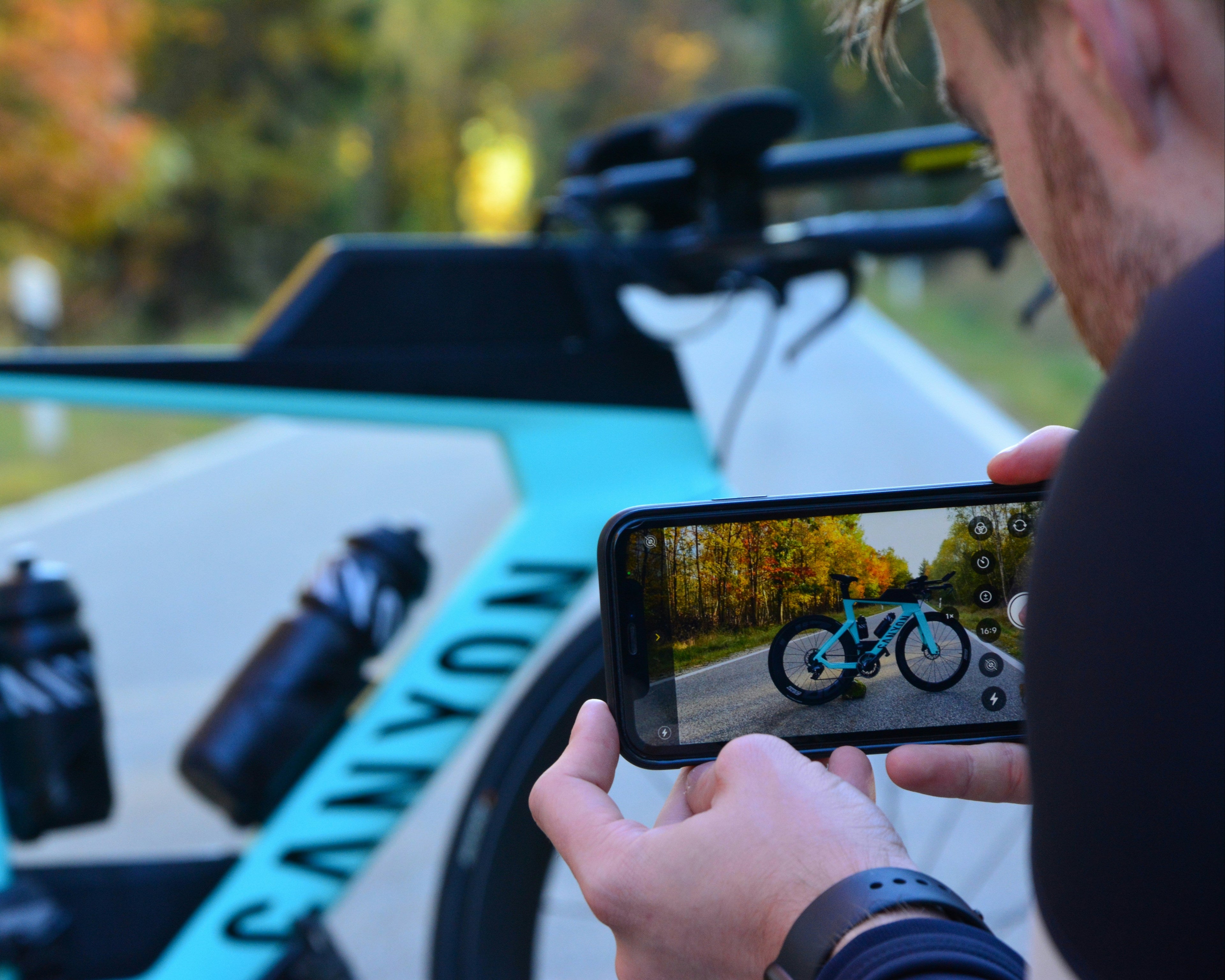 A person is taking a picture of a bicycle with a smartphone. The bicycle is mounted on a car's roof rack, and the brand 'CANYON' is visible on the bike and the car.